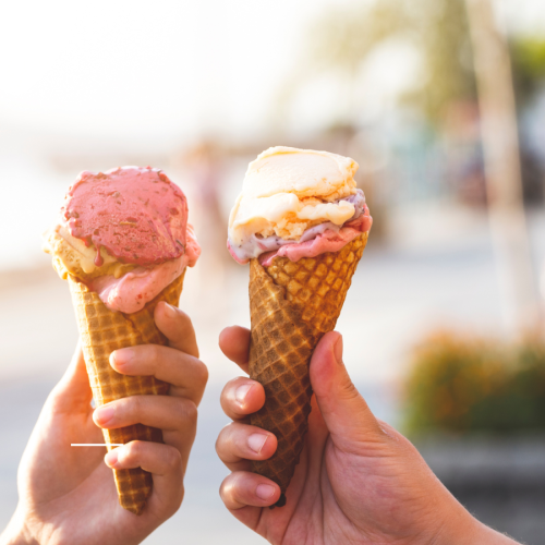 Two hands hold ice cream cones with pink and white scoops melting slightly. A sunny, blurred Key West street scene forms the background, suggesting a warm outdoor setting.