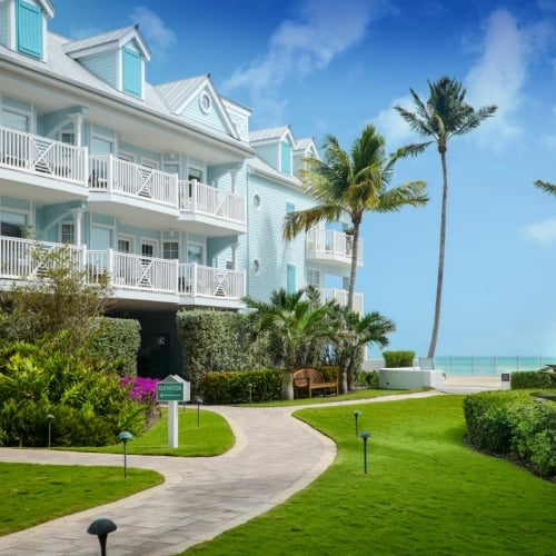 A pastel blue building with white balconies stands amidst lush gardens and palm trees near a turquoise ocean in Key West. A sign reads