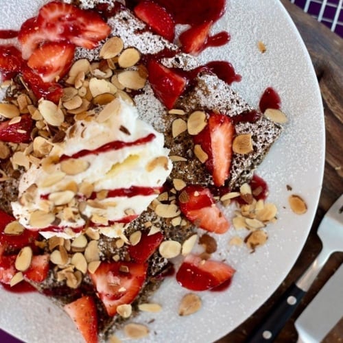 A plate of crepes topped with whipped cream, sliced strawberries, almonds, and berry sauce, resting on a wooden board in Key West. A fork and knife are placed beside it.