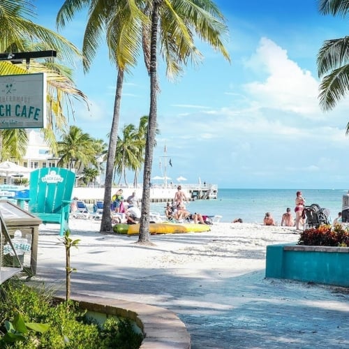 Colorful totem poles stand in a courtyard, adorned with patterns and bird sculptures. A person in a yellow dress sits beside them, with a striped wall and palm trees in the background. Key West.