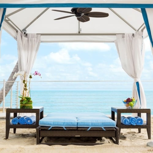 A beach cabana in Key West features a cushioned lounge with rolled towels, flower and fruit arrangements, surrounded by white curtains under a fan, with a view of the ocean.