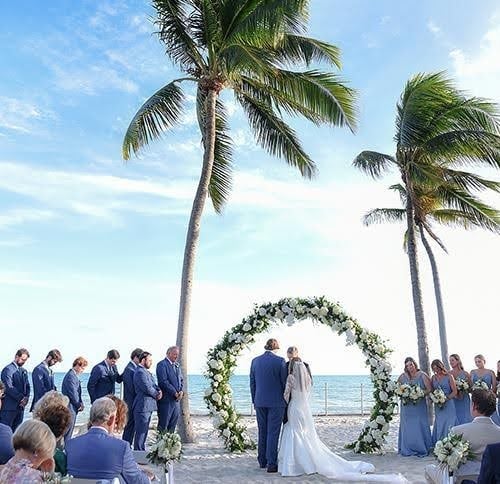 A couple stands under a floral arch exchanging vows, surrounded by their wedding party on a sandy beach with palm trees, against a backdrop of blue sky and ocean in Key West.