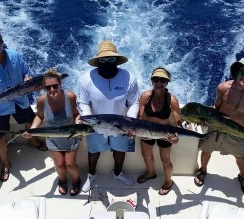 Five people stand on a boat holding freshly caught fish, smiling under the sun, with ocean waves trailing behind. The scene is set off Key West or The Keys.