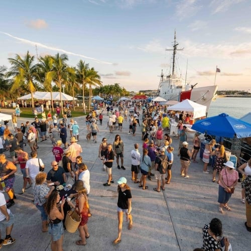 A lively crowd mingling at a waterfront festival alongside a docked ship, with palm trees and various vendor tents under a clear sky in Key West.