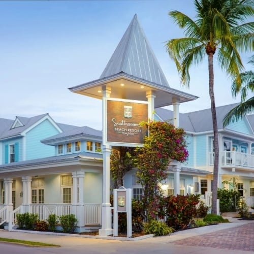 A pastel blue and white beach resort with colonial architecture stands among palm trees. A sign reads