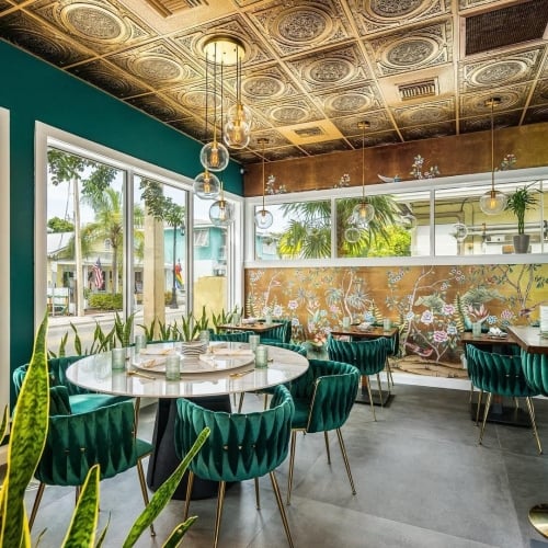 Elegant dining room featuring a round table with green velvet chairs surrounded by lush plants. Ornate ceiling and floral wall mural in Key West create a luxurious ambiance.