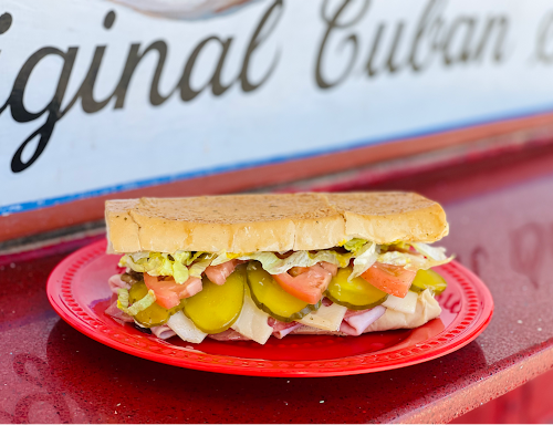 A Cuban sandwich rests on a red plate, displayed on a counter in Key West. The sandwich is filled with sliced pickles, tomato, lettuce, and meats. A sign reads