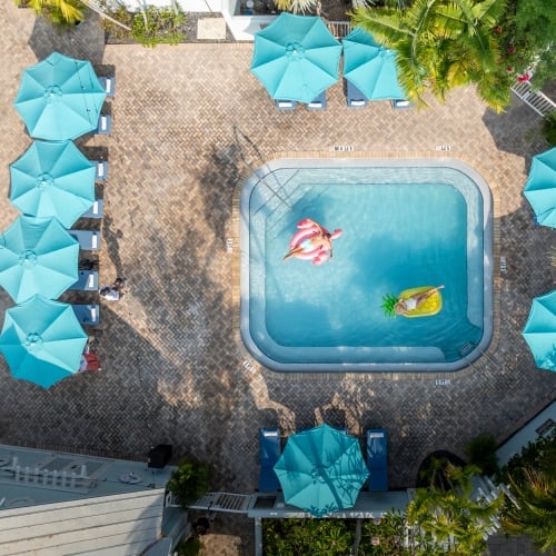 Two people float on inflatable toys in a pool surrounded by teal umbrellas and lounge chairs on a brick patio. Palm trees enhance the tropical setting of The Keys.