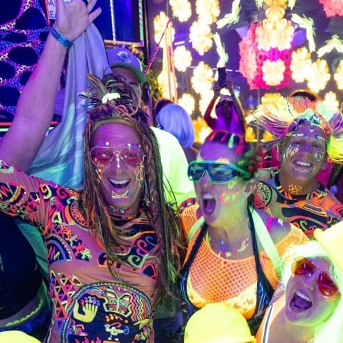 People in colorful, neon face paint and clothing, smiling and dancing energetically under UV lighting. The vibrant party atmosphere includes glowing decorations and joyful expressions, set in Key West.