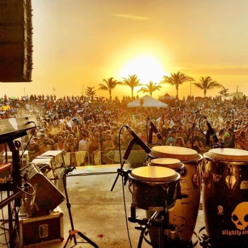 Congas and a keyboard on a stage overlook a large crowd at an outdoor concert during sunset in Key West. Text: