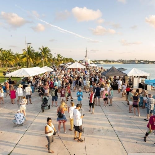 Crowd enjoying a waterfront festival with tents, palm trees, and a clear sky in Key West. Text on blue tent:
