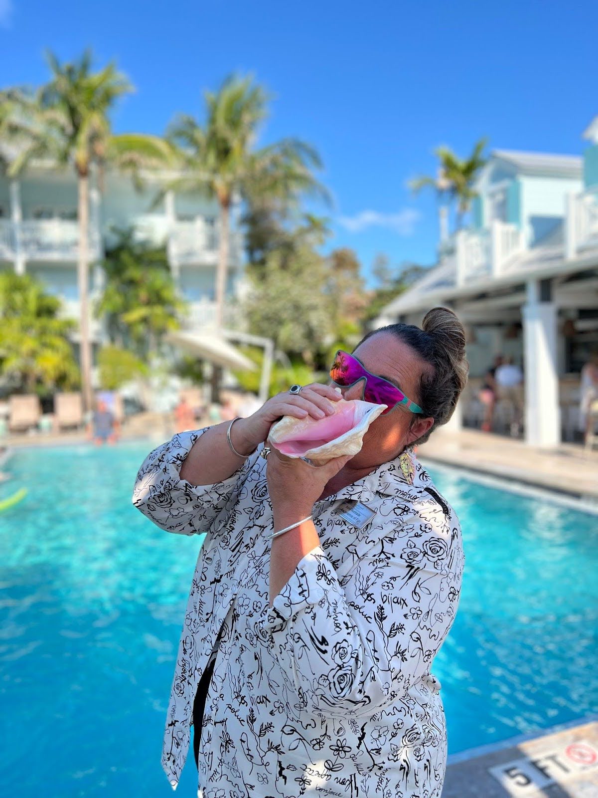 A woman standing poolside blows into a conch shell.