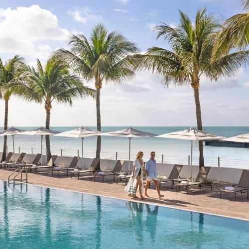 Vista of a pool with palm trees and the ocean in the background. A couple walks along the pool.
