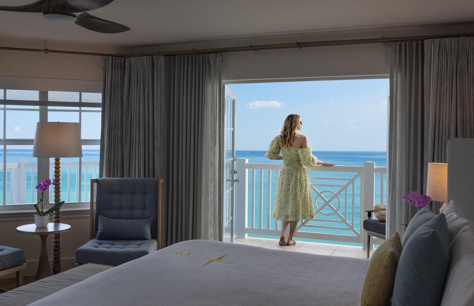 A woman stands on her balcony looking out over the water on the blue sky day. 