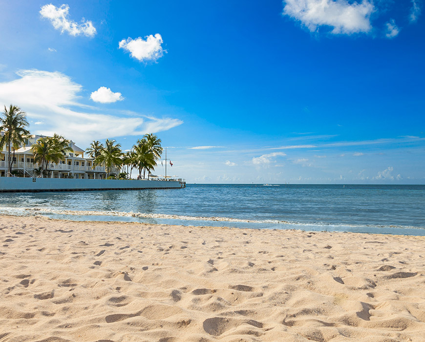 A beach with a white building and palm trees in the background, facing the ocean under a blue sky.