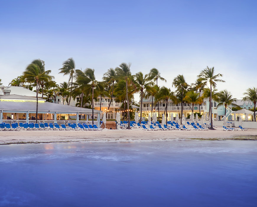 A beachfront view of a resort with palm trees and blue beach chairs