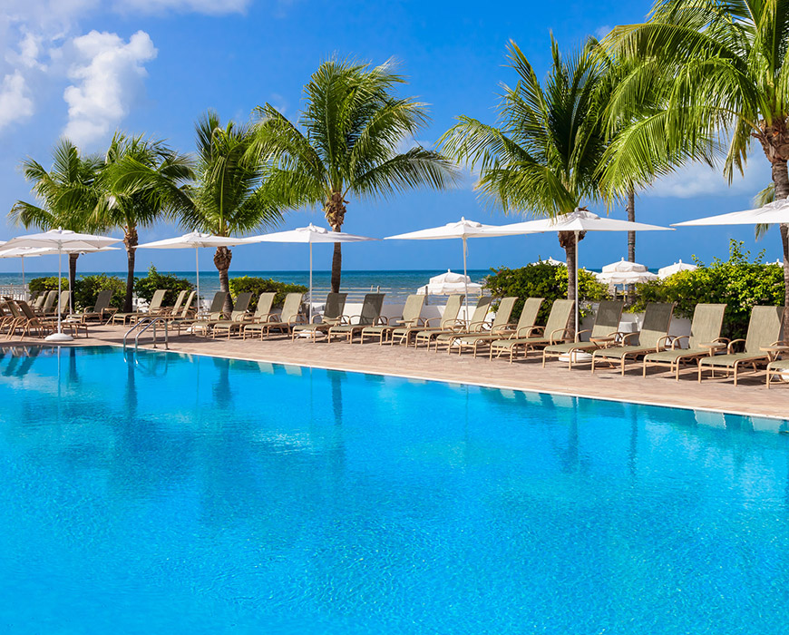 A serene pool scene with lounge chairs, white umbrellas, and palm trees overlooking the ocean.
