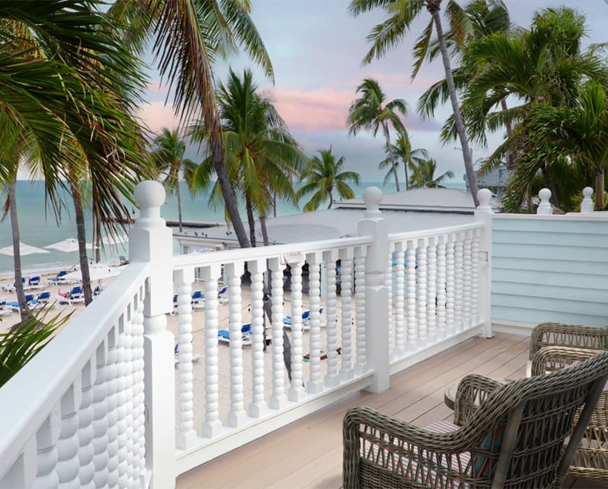 A balcony with a white railing and wicker chairs overlooking a beach with palm trees and an ocean view at sunset.