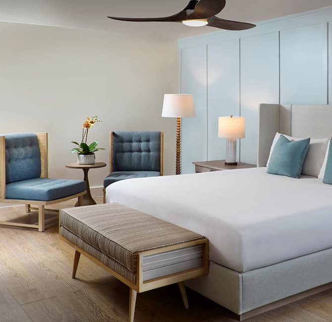 A hotel room with a large bed, two blue chairs, and a ceiling fan.