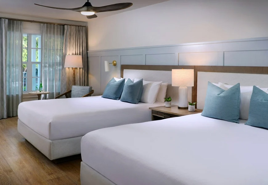 A hotel room with two queen beds, white bedding, and blue accent pillows.