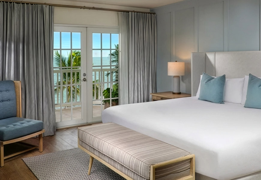 A hotel room with a king-sized bed, balcony access, and a view of the ocean and palm trees.