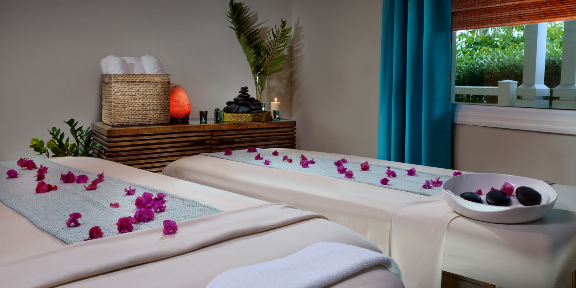 Two massage beds with white sheets and towels, stones, and flowers in a spa room with a window.