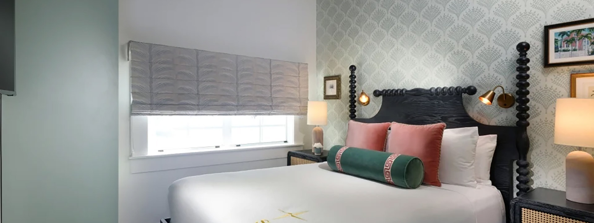 A neatly made bed with pillows and a bolster in a room with a window, nightstand, and lamp.