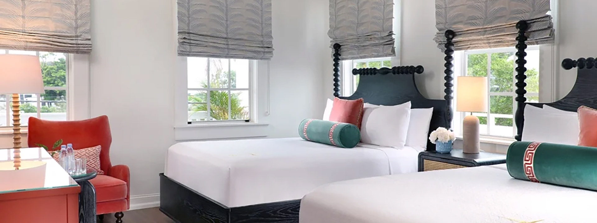 A brightly lit hotel room with two beds, a red chair, and a table with a lamp.