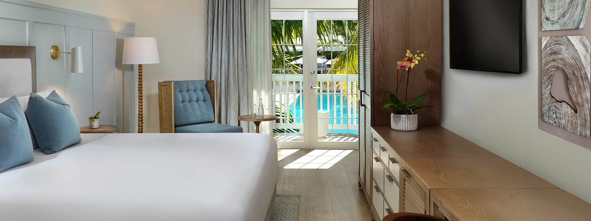 A hotel room with a king-sized bed, dresser, TV, and balcony overlooking the pool.