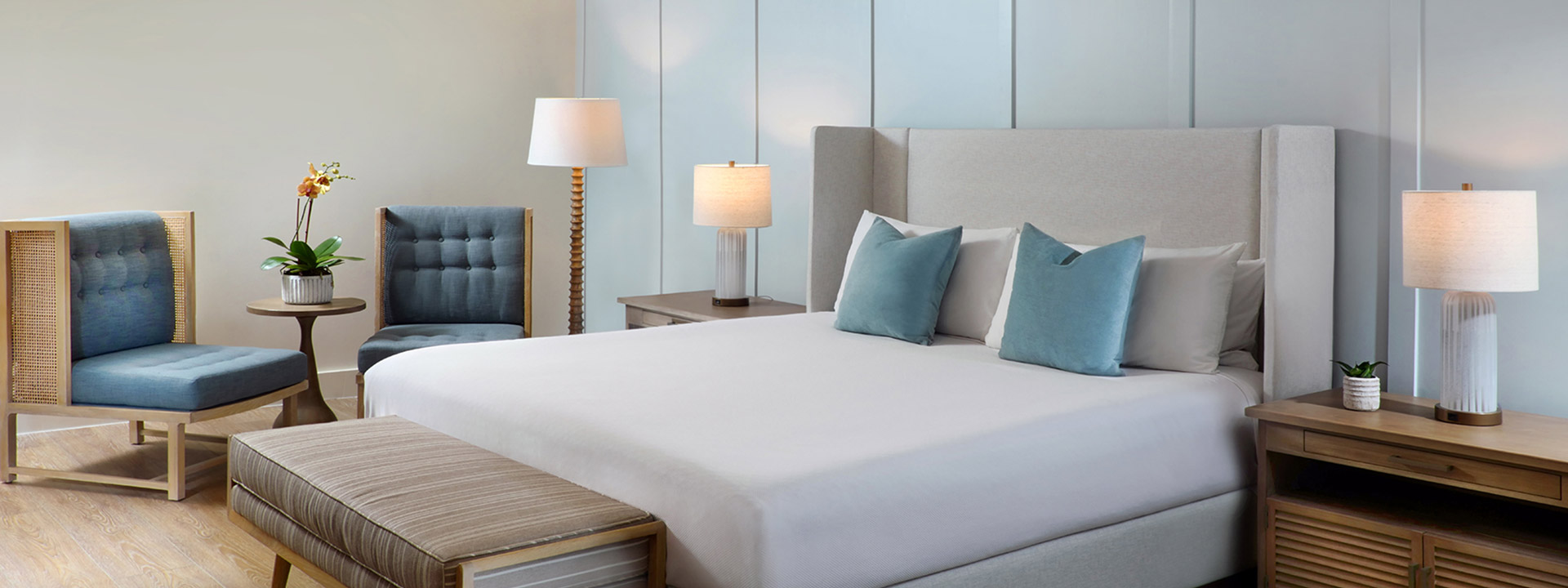A neatly made king-sized bed with blue and white pillows, flanked by two nightstands with lamps, in a room with light wood flooring and blue accents.