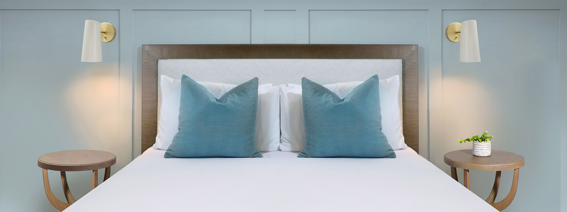 A neatly made bed with white bedding, blue pillows, and a wooden headboard, flanked by round nightstands with wall lamps.